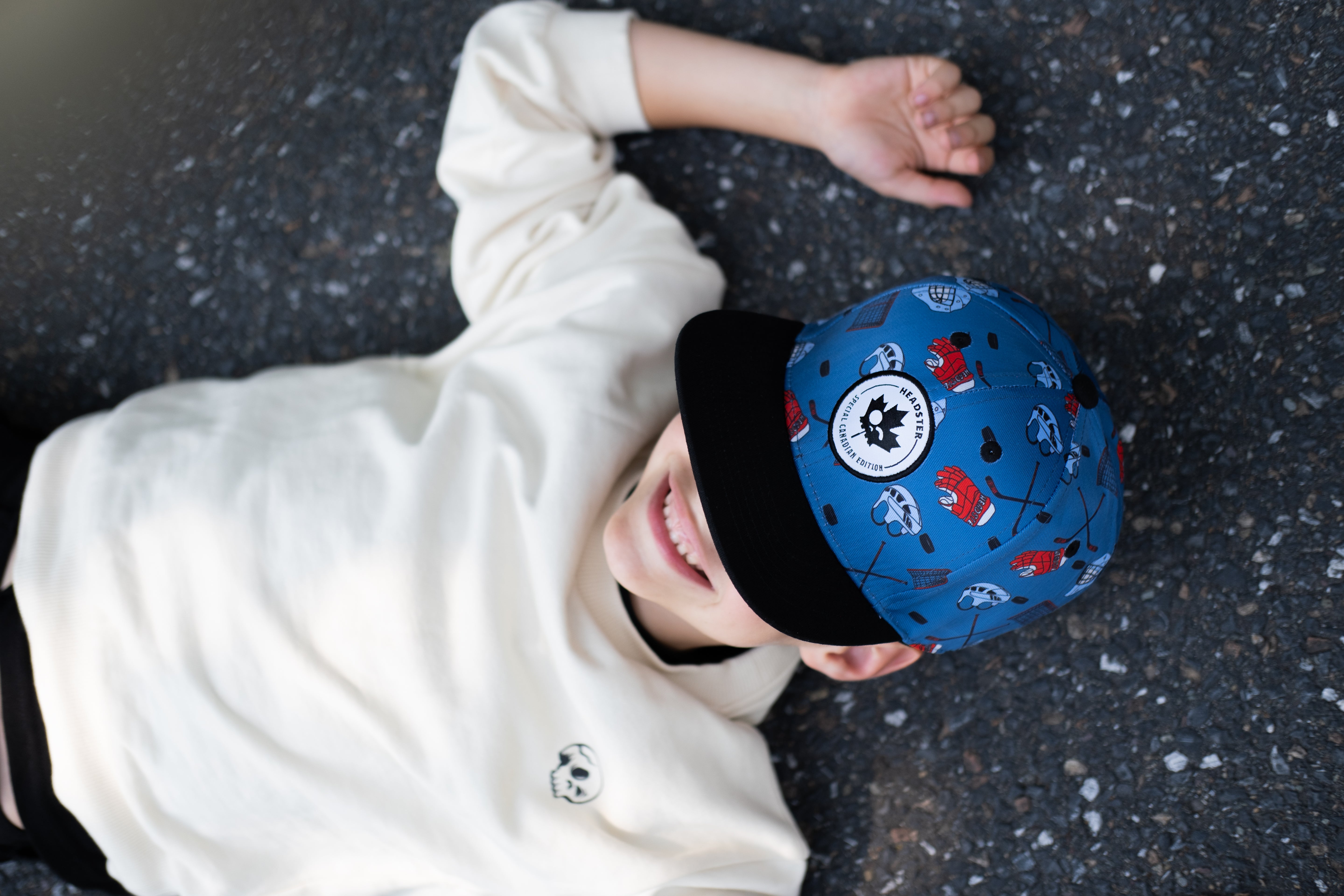 A child lying on asphalt wearing a blue patterned baseball cap with a black brim and a cream sweatshirt, photographed from above.